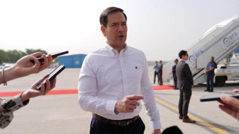 Marco Rubio, wearing a white shirt and dark trousers, on the tarmac of an airport, point with his finger as he speaks to journalists - not in shot, just a couple of hands with phones recording him. Other people can be seen in the background, near the stairs of an airplane.