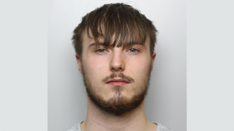 A police mugshot of Ben Churcher. He is wearing a grey t-shirt and standing against a grey background. He has blue eyes, dark brown hair which is styled downwards over his forehead, and a dark beard and moustache. He is looking at the camera with a blank expression. 