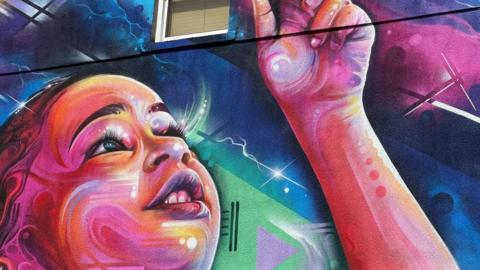 A colourful paint drawing of a young girl on the side of a wall with a window near the top centre. 