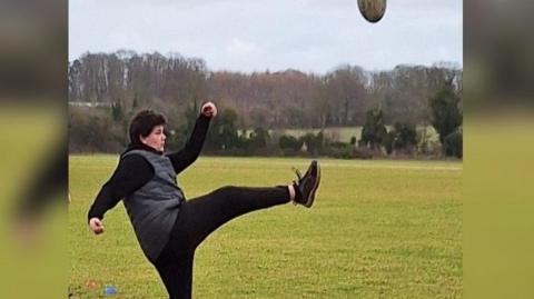 Student Isaish in black trousers, top and trainers kicking a rugby ball on a field.