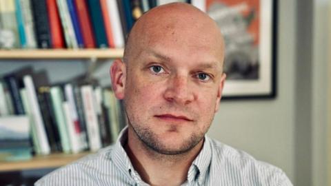 A middle-aged man in a light grey and white striped shirt sits in front of a bookcase looking directly into the camera 