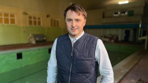 A man with dark hair and wearing a striped shirt and navy gilet, stands in front of an empty swimming pool