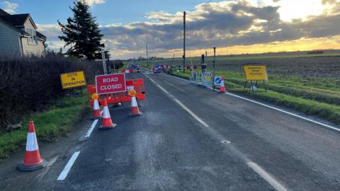 B1049, Twenty Pence Road, very rural setting, one house on the left is the only property visible, farmland is left and right of the roadway, various signs around an electronic barrier that can only be used by residents with a special key fob, other drivers can't use it.