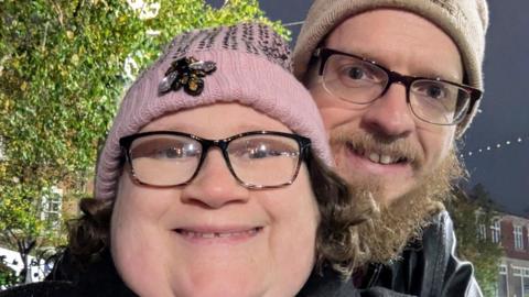A woman with black rimmed glasses and a pink bobble hat smiles at the camera. Behind her a man with a beige bobble hat cuddles up to her. He has black rimmed glasses too and a dark beard.