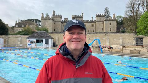 A middle-aged man wearing a grey and red coat and a dark cap with a white emblem on smiles as he stands in front of a pool with lanes. There is a large, castle-like stately home on the other side of the wall.