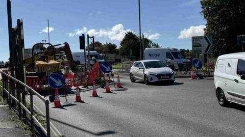 A road with orange traffic cones cones, a yellow digger, a worker in high-vis clothing and a white hard hat, and two blue signs with white arrows. There are three white vans and a white car in the road.