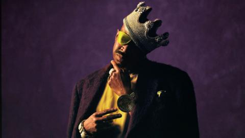 Slick Rick wears a crown and a golden eyepatch, while standing against a regal purple background, in a promotional photo for his 2025 album, Victory
