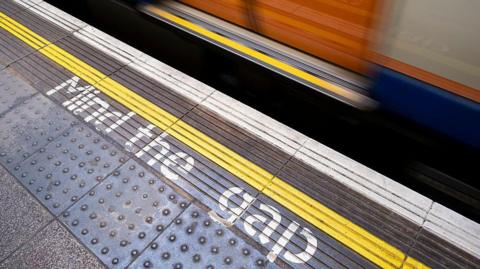 Close-up view of a Tube station platform featuring a yellow line and the words "Mind the gap" printed in white letters. The bottom of some train doors can also be seen.