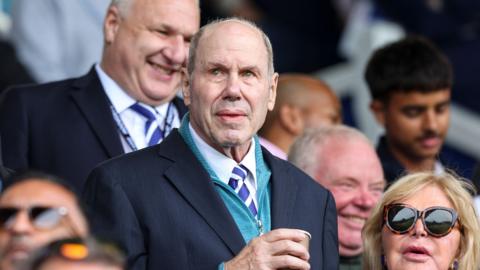 A close-up of Michael Eisner, wearing a blue jacket with a light blue jumper, white shirt and blue and white striped tie, amongst the crowd at a Portsmouth game
