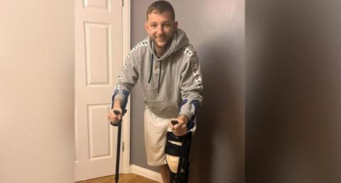 Joe Collins holding crutches with one leg in a black brace. He has short dark hair and has stubble, and is wearing a grey Nike hoodie and white shorts. He is standing indoors with a grey wall and white door behind him. 