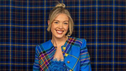 A blonde haired woman wearing a blue tartan jacket against a blue tartan backdrop