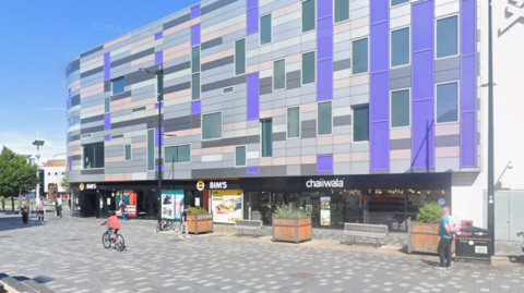 Luton town centre: A grey, blue and pink rectangular building overlooking a paved area. Three square wooden planters stand on the paved area. Two people are walking in front of the building and one is riding a cycle.