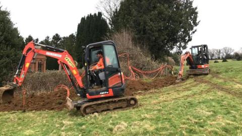 Two small orange diggers digging up grassland. beside them is orange cabling hanging from a hedge