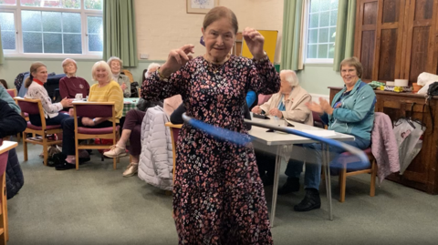 Nova Strange twirling a hula hoop around her while people watch seated in the background. She has tied-back brown hair and is wearing a long floral dress.