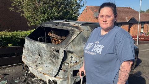 Leonie McLaughlin holds a walking stick as she stands in front of a burned out car. She is wearing glasses and a grey/blue t shirt.