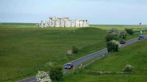 A303 with cars travelling along with Stonehenge in the distance. You can also see large fields on either side.