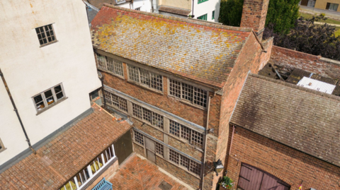 A drone shot of an old pin factory in Gloucester. It is a brick building that sits in a courtyard and has three rows of windows with wooden frames