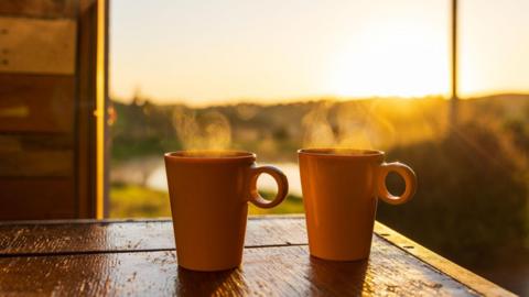 Two orange mugs are set on a wooden table with steam rising from them.
