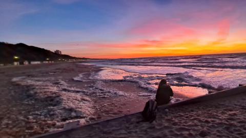 A solitary person sits on a beach with their backpack resting beside them. The waves are crashing on the sand in front of them while the sky bursts full of orange, yellow, purple and blue hues form a stunning sunset. 