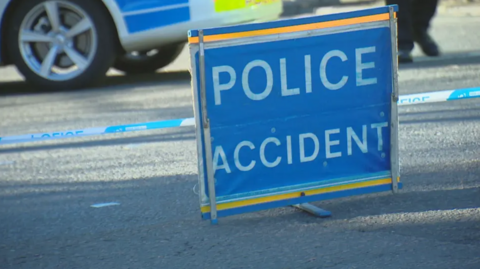 A sign can be seen which is blue and has white font which reads 'POLICE ACCIDENT'. Behind the sign there is police tape and the bottom of a police car. 