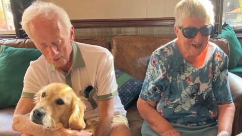 Jeff and Janie Johnson and one of their guide dogs. Jeff wears a white T-shirt and shorts, hugging a golden retriever guide dog, Janie is dressed in a blue and pink skirt and blouse, and is wearing dark glasses