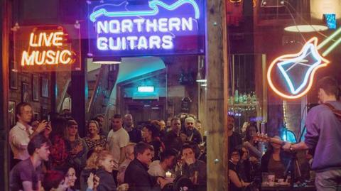 The outside of Northern Guitars music venue in Leeds is pictured. There are three neon signs in the window - one saying 'live music', one saying 'Northern Guitars' and one with the silhouette of a guitar. A man is playing the guitar and singing, as a crowd looks on.