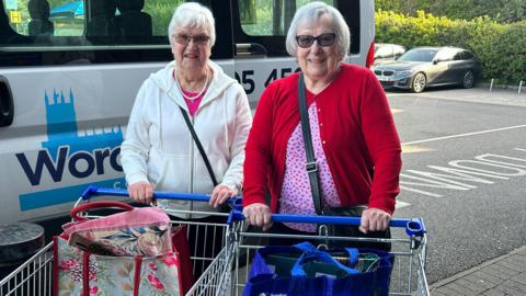 Two elderly women wearing tinted glasses stand in front of a white minibus, their hands on shopping trolleys with shopping bags in them. One has short white hair and is wearing a cream-white hoodie over a pink top. The other has a white bob and is wearing a red cardigan over a pink and red top.
