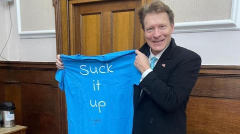 Richard Tice MP, the national party deputy leader of Reform UK, pictured holding up a T-shirt with Suck It Up written on it in white lettering.