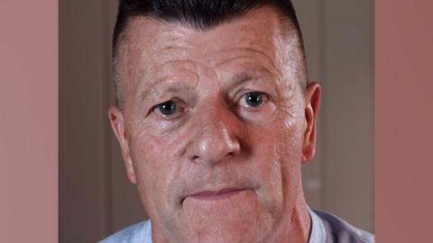 A man with dark cropped hair looks down the lens with a serious expression 
