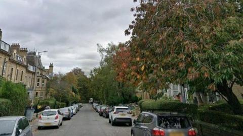 A Victorian terraced street lined with trees and parked cars. It is a cloudy day.