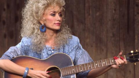 Dolly Parton strums a guitar while looking to the right of frame wearing a blue top against a background of wooden panels on ABC TV in 1987.