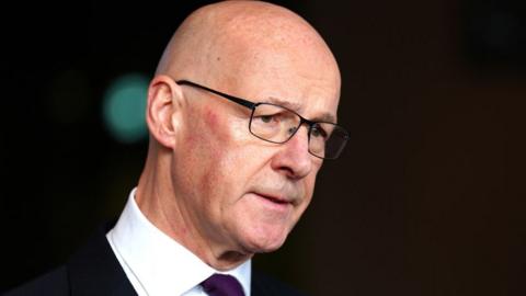 John Swinney, a bald man wearing glasses and a dark suit with a white shirt, speaks to the media in front of an out of focus black background 