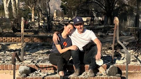 Harry Butler is pictured sitting on steps at the front of his destroyed home. Pictured next to him, with her head on his shoulder, is his fiancée Vanessa. Rubble and burned trees can be seen in the background.