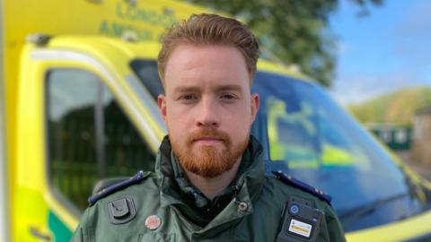 James is standing in front of a fluorescent yellow ambulance. He has light brown hair and a beard. He is wearing a dark green ambulance service jacket with a body-worn camera. 