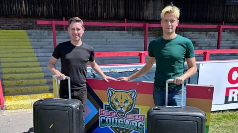 Ryan and Kaue stand on the rugby pitch in front of a banner for the club which is adorned with the club's logo against a rainbow flag. Ryan has black hair whilst Kaue has bleach blonde hair. They are holding suitcases