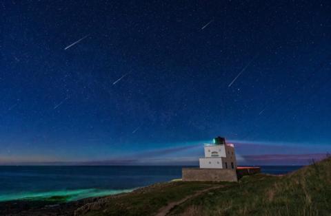 Meteors over a lighthouse at Bamburgh, perched on a grassy bank. The light in the sky is blue, with purple and green tones.