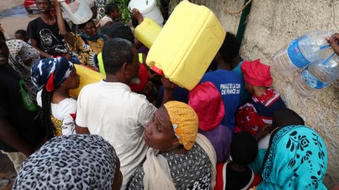 A crowd of people queue for water in Mayotte, some hold large water containers above their heads