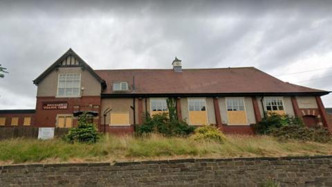 A large red-brick building, some of its windows are boarded up, a sign says Brierley Village Club.