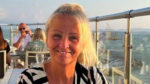 Anita Rose smiling into the camera. Her blonde hair is in a pony tail and she is sat at a rooftop restaurant is wearing a black and white striped top. Mountains/hills are visible on the horizon.