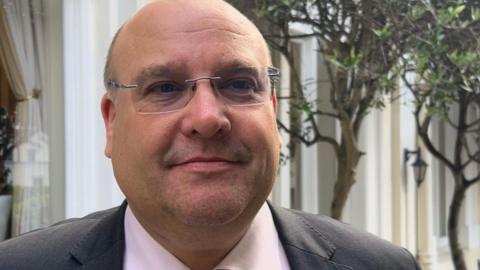 A bald man with rectangular glasses smiles at the camera in front of a hotel courtyard, with spindly green trees and trestle tables behind him. he is wearing a grey suit, with a white shirt and red tie.