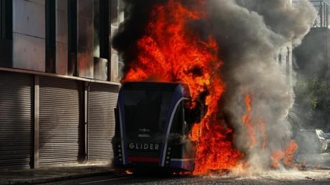 A bus has been set on fire. There are bright orange flames and thick grey and black smoke coming from the bus