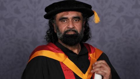 A man with long black curly hair and a black and grey beard is wearing a graduation gown, with gold and red colours, and holding a rolled decree