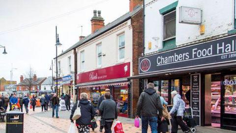 Shoppers in Arnold town centre carrying bags. There is a pedestrianised area in the foreground and a row of shops in the background.