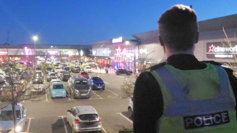 A police officer stands with his back to us. He is watching over the car park on a retail park, late at night.