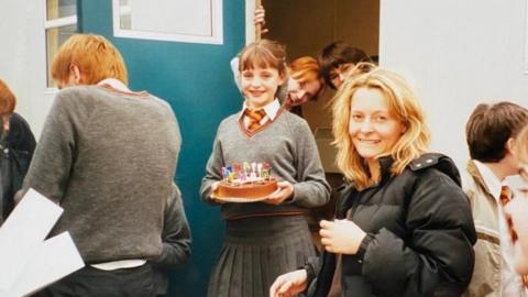 A young girl dressed in a Hogwarts school uniform is smiling holding a birthday cake. She is surrounded by people, some resembling characters from the Harry Potter movies.