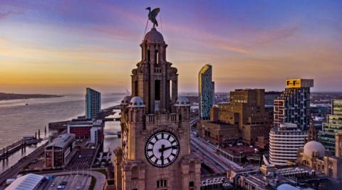 A view of the clock and Liverbird on the Liverpool Liver building with the River Mersey in the background taken at sunrise. A number of high-rise buildings are in the background.