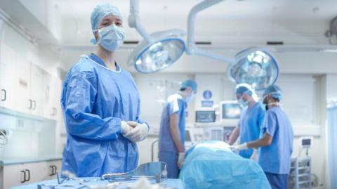 A nurse dressed head to toe in blue scrubs including a blue face mask and hair net. She is standing in front of a table with surgical equipment on it, inside an operating theatre. In the background, a patient lies on a bed under a blue blanket, with three other healthcare workers around them, also in scrubs.