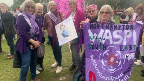 Jill Robertson, who has blonde hair and glasses, holds up a purple banner with "Northampton Waspi" written on it. She is on the right, standing at the end of a line of five other campaigners, who are also looking at the camera. The picture has been taken at a protest at Westminster.