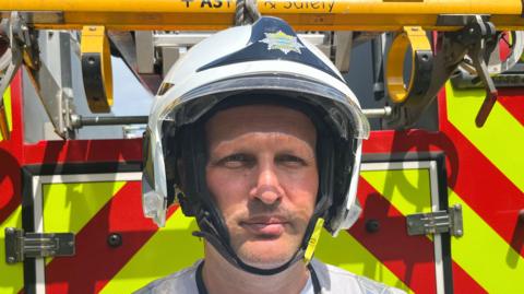 A firefighter wearing his uniform, looking straight at the camera standing at the back of a fire engine.