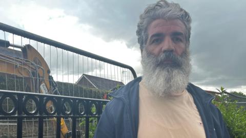 A man standing next to a metal railing fence with a digger working behind him. He has grey hair and a grey beard, and wearing a beige top and a blue jacket